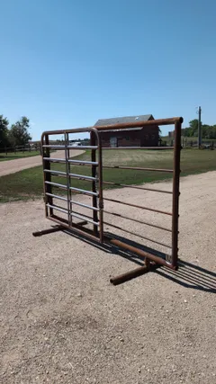 Free-standing pipe panel with a 180-degree swinging walk-through gate and chain latch, standing on its skid feet in a farmyard